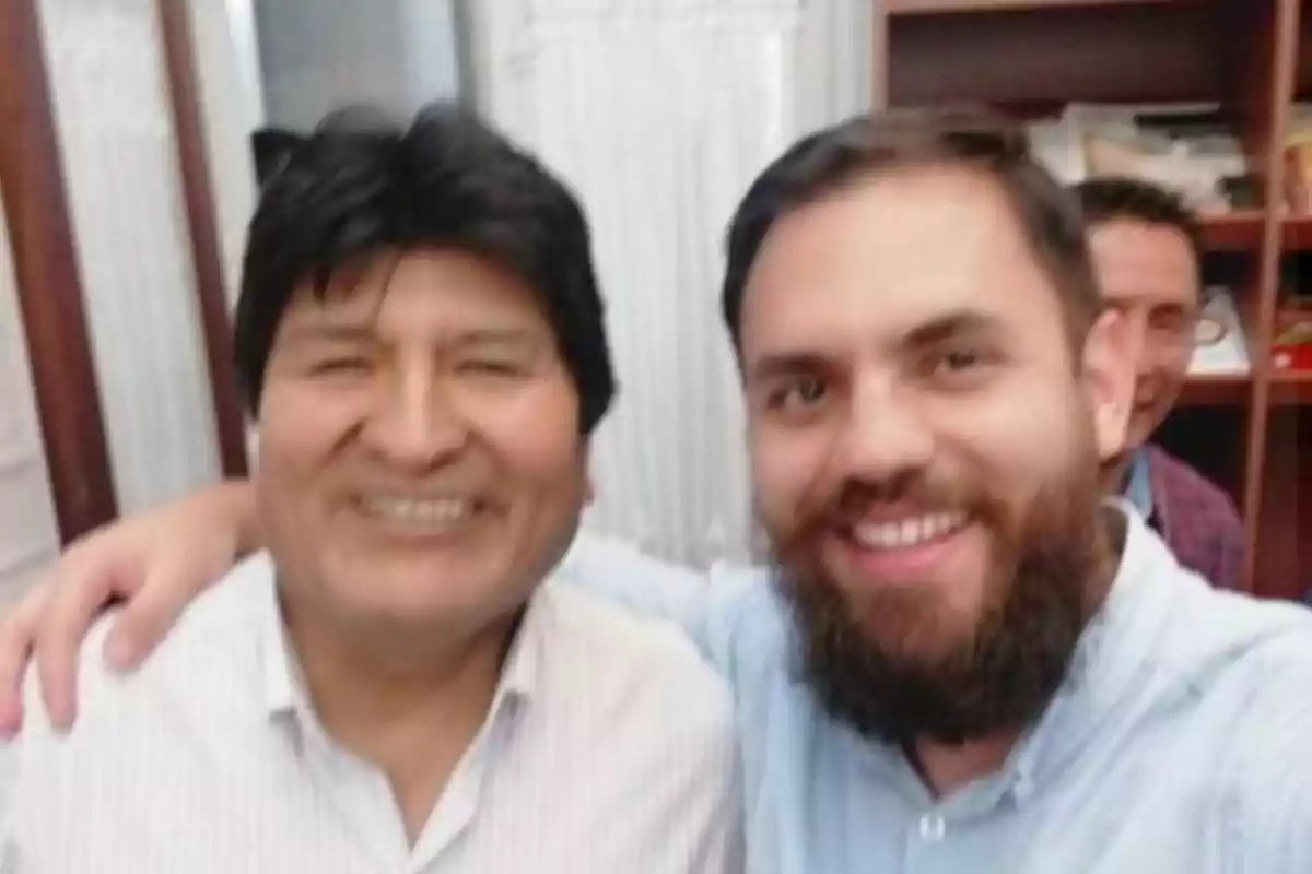 Two men smile while taking a selfie indoors, with another person in the background.