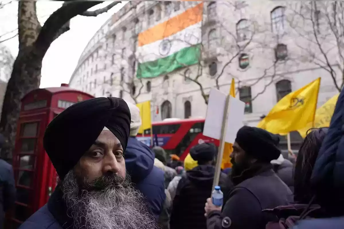 Un hombre con turbante en una manifestación frente a un edificio con una bandera de India y una cabina telefónica roja.