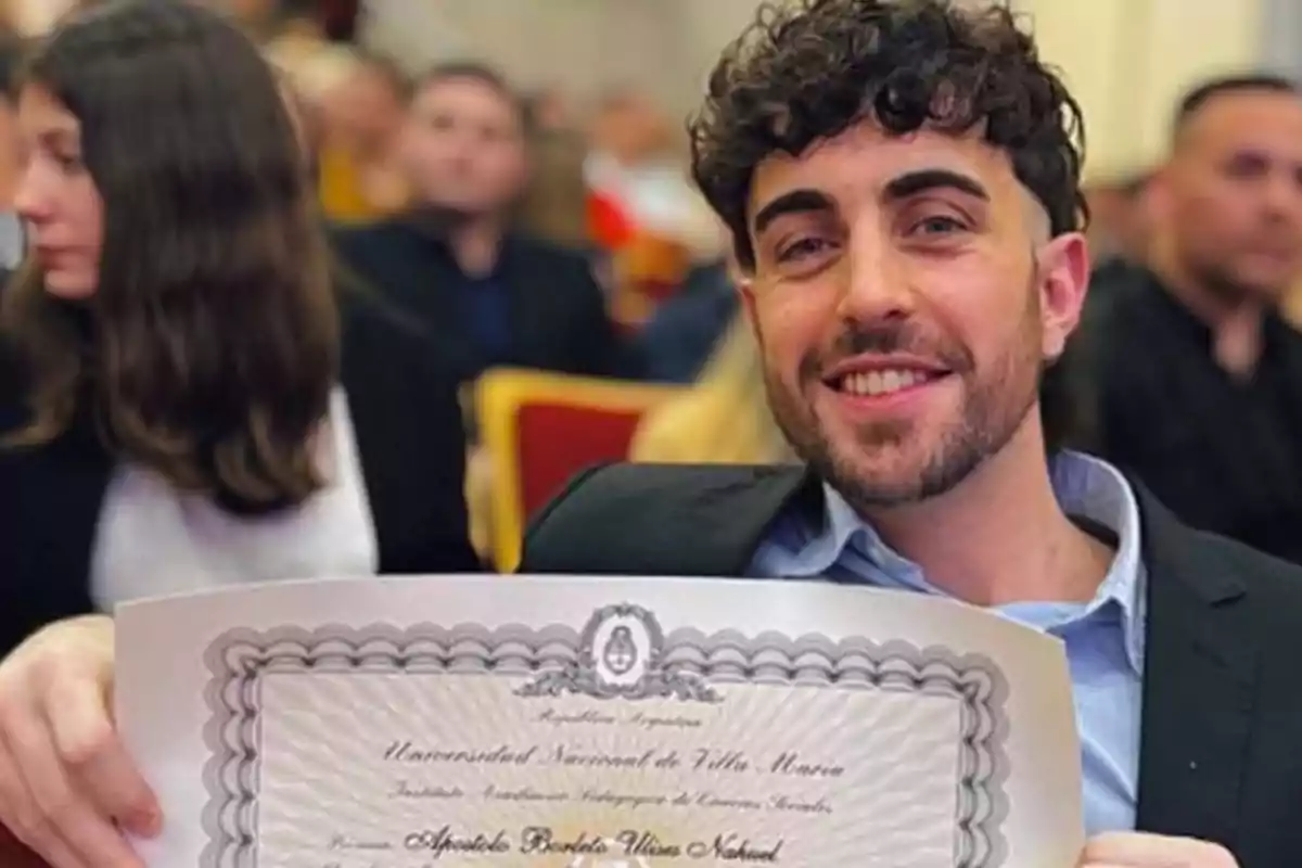 A smiling man holds a diploma at an event with several people sitting in the background.