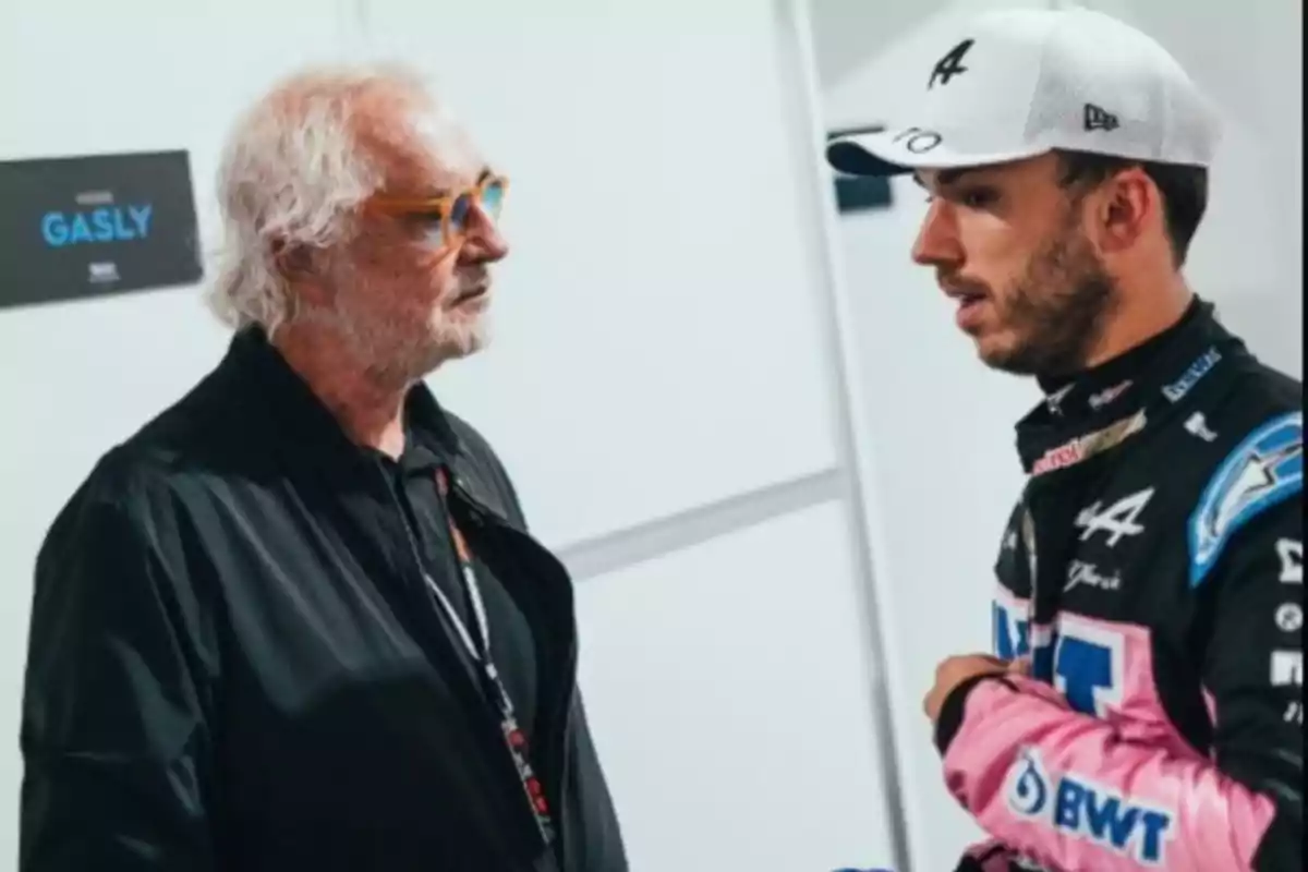 Two men are talking in a hallway; one is wearing glasses and a black jacket, the other is dressed in a racing uniform and a white cap. Two men are talking in a hallway; one is wearing glasses and a black jacket, the other is dressed in a racing uniform and a white cap.