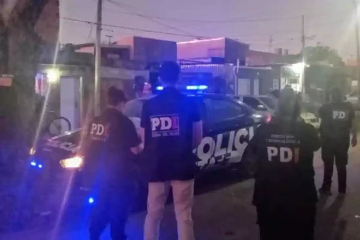 Personas con chalecos de la PDI junto a un vehículo policial con luces encendidas en una calle por la noche. Personas con chalecos de la PDI junto a un vehículo policial con luces encendidas en una calle por la noche.