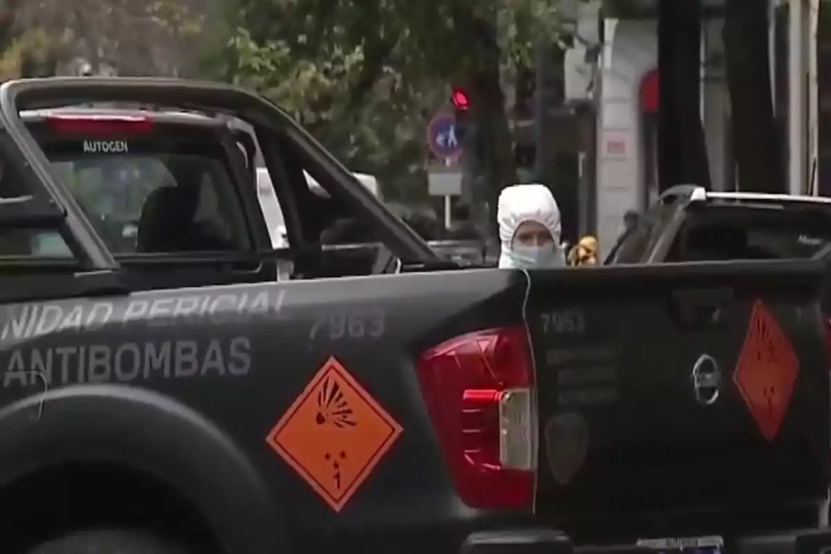 Una camioneta de la unidad pericial antibombas con señales de advertencia y una persona con traje de protección en el fondo. Una camioneta de la unidad pericial antibombas con señales de advertencia y una persona con traje de protección en el fondo.