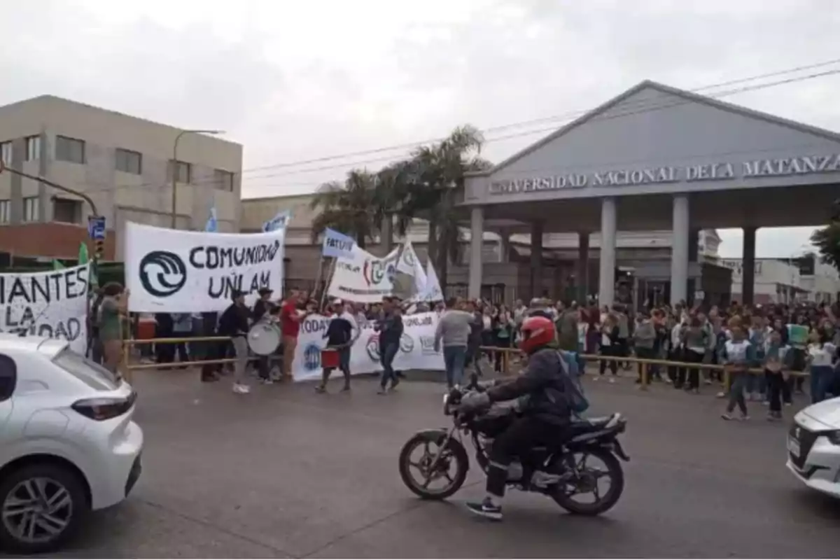 Una multitud se reúne frente a la entrada de la Universidad Nacional de La Matanza, sosteniendo pancartas y banderas, mientras un motociclista pasa por la calle.