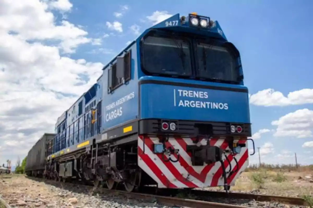 Un tren de carga azul de Trenes Argentinos circula por las vías bajo un cielo parcialmente nublado.