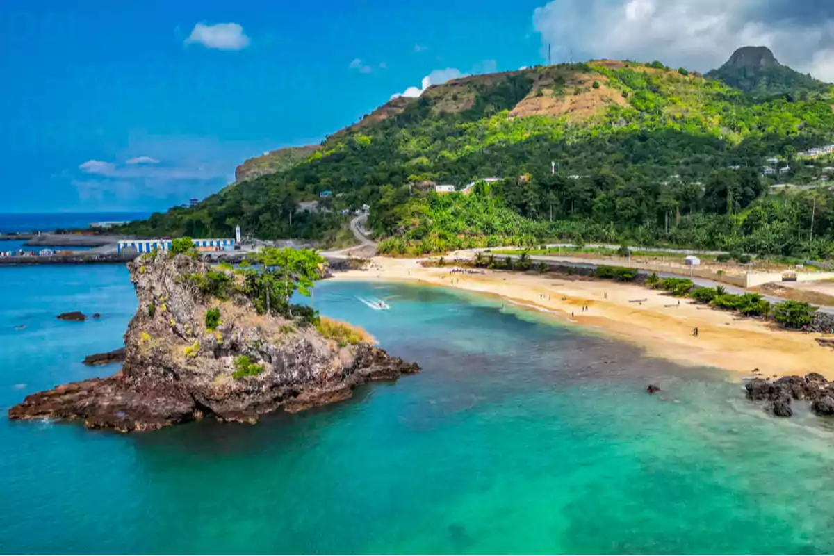 Golden sandy beach with turquoise waters and a rocky formation covered in vegetation facing a green hill under a blue sky Golden sandy beach with turquoise waters and a rocky formation covered in vegetation facing a green hill under a blue sky