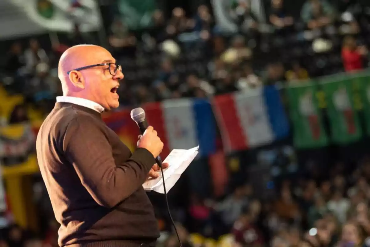 Bald man with glasses speaking into a microphone and holding papers in front of a crowd at an event with flags in the background Bald man with glasses speaking into a microphone and holding papers in front of a crowd at an event with flags in the background