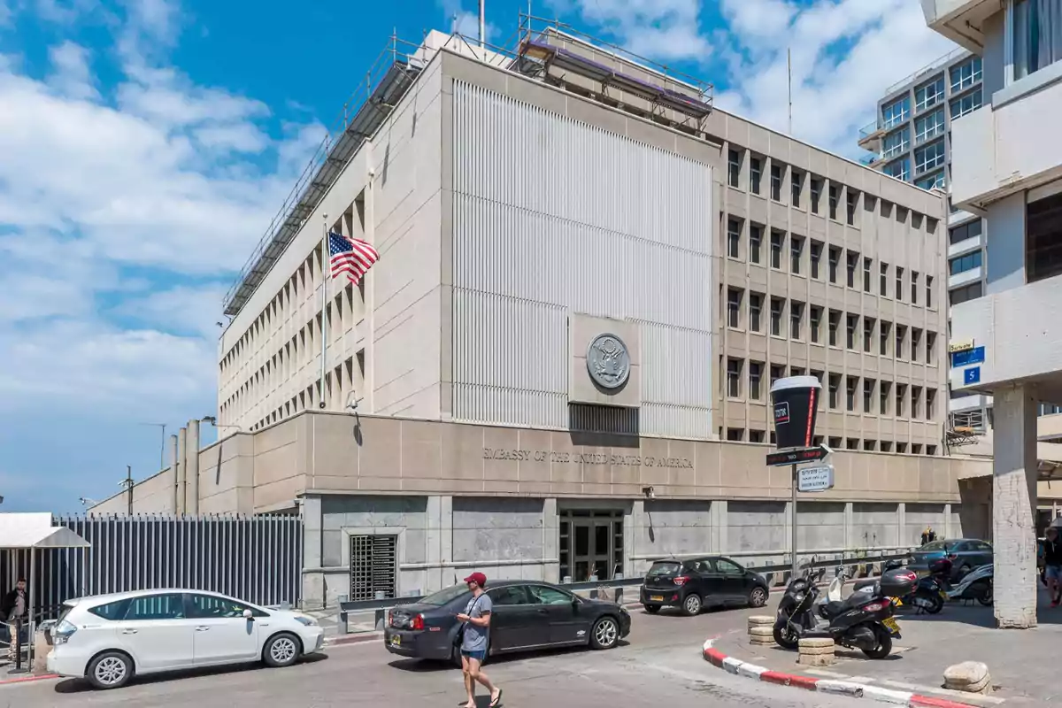 Edificio de la embajada de Estados Unidos con una bandera estadounidense ondeando al frente y varios autos estacionados en la calle