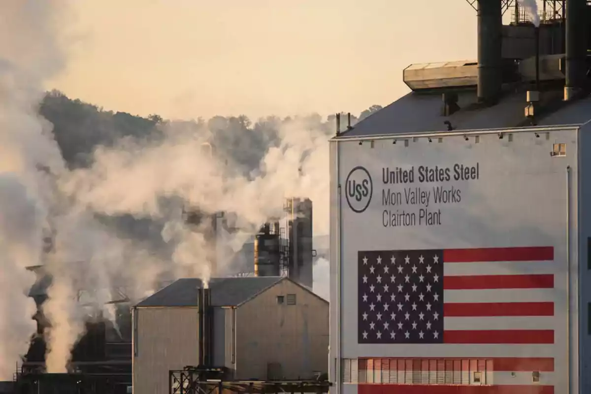 Fábrica de acero con humo saliendo de las chimeneas y una bandera de Estados Unidos en el edificio.