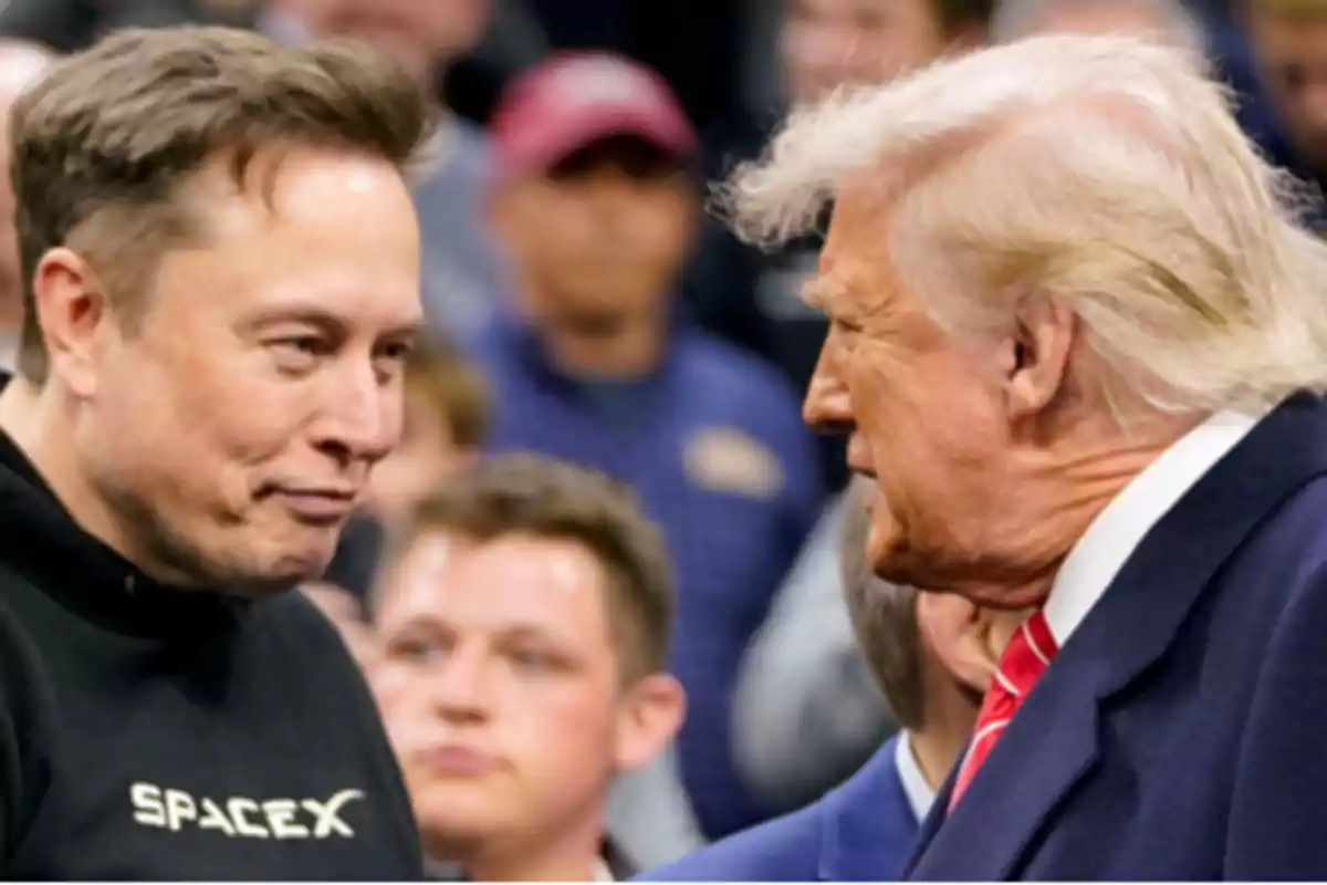 Dos hombres conversan cara a cara en un evento rodeados de personas, uno lleva una sudadera negra con el logo de SpaceX y el otro viste traje azul con corbata roja Dos hombres conversan cara a cara en un evento rodeados de personas, uno lleva una sudadera negra con el logo de SpaceX y el otro viste traje azul con corbata roja