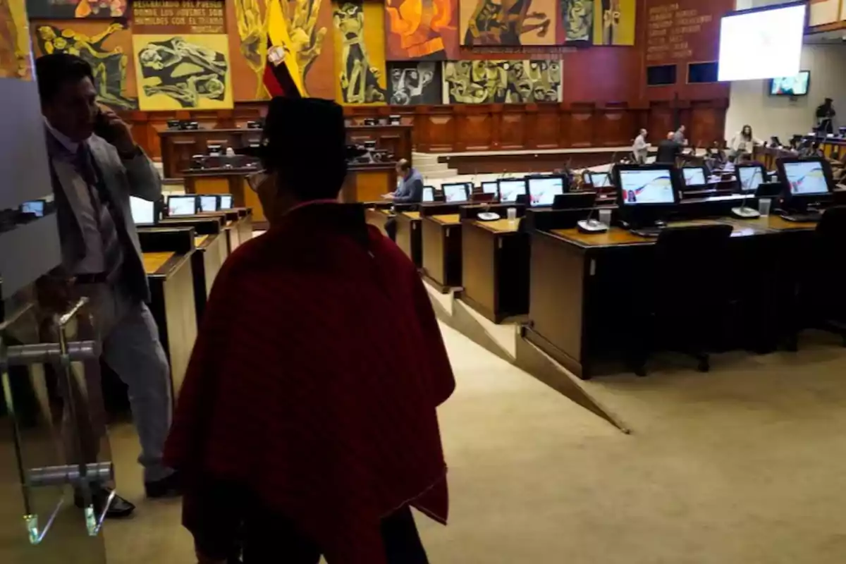 A person in traditional attire walks inside a legislative building with colorful murals on the walls and several workstations with computers.