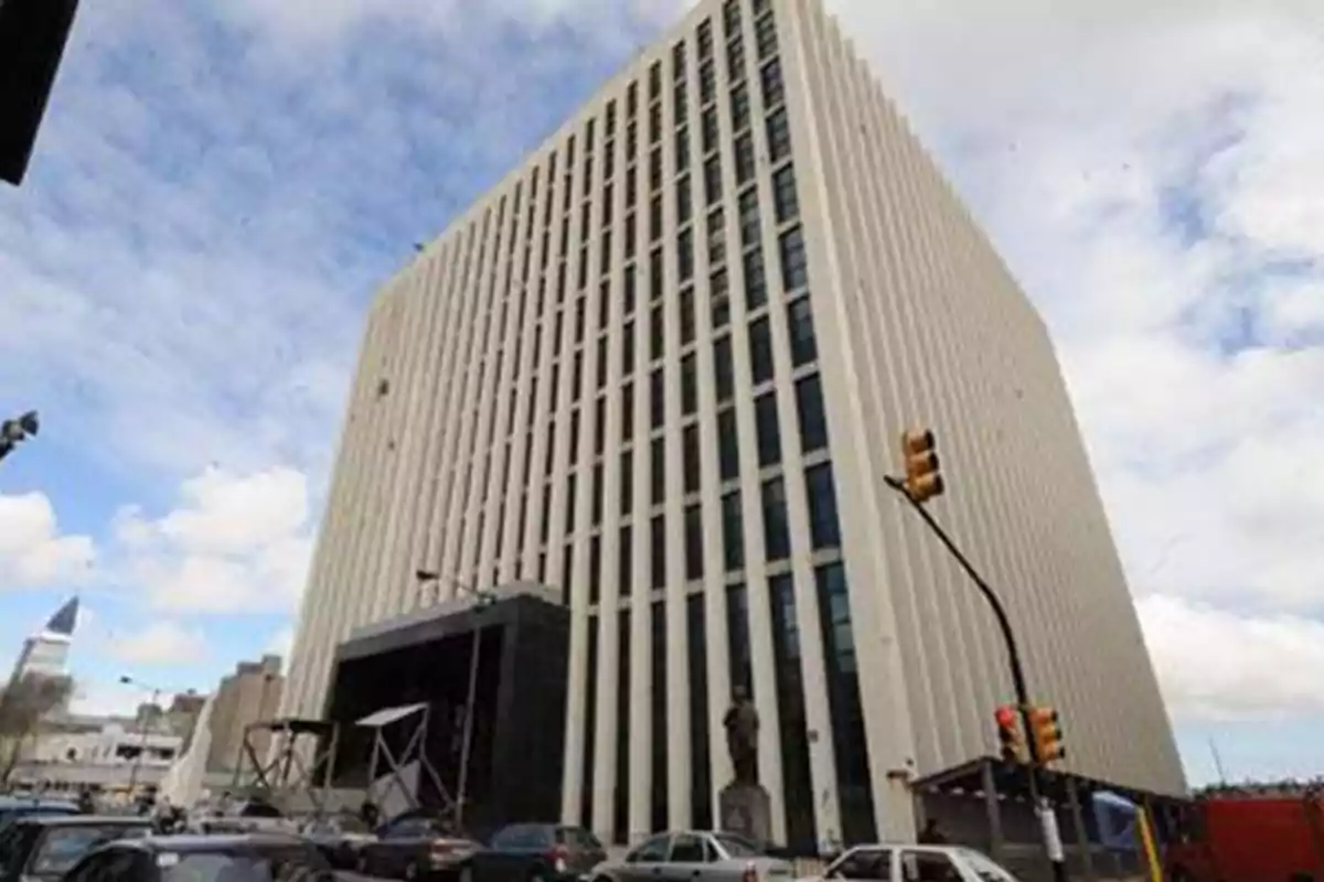 Un edificio alto con fachada de columnas verticales y ventanas oscuras bajo un cielo parcialmente nublado con tráfico y semáforos en la calle. Un edificio alto con fachada de columnas verticales y ventanas oscuras bajo un cielo parcialmente nublado con tráfico y semáforos en la calle.
