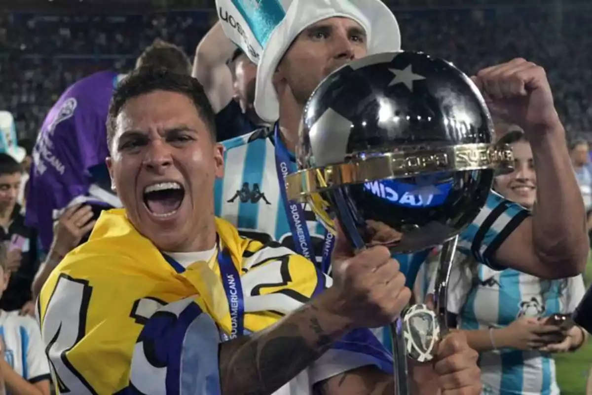 Player celebrating enthusiastically with a soccer trophy while wearing a flag over his shoulders, surrounded by teammates and fans in the stadium. Player celebrating enthusiastically with a soccer trophy while wearing a flag over his shoulders, surrounded by teammates and fans in the stadium.