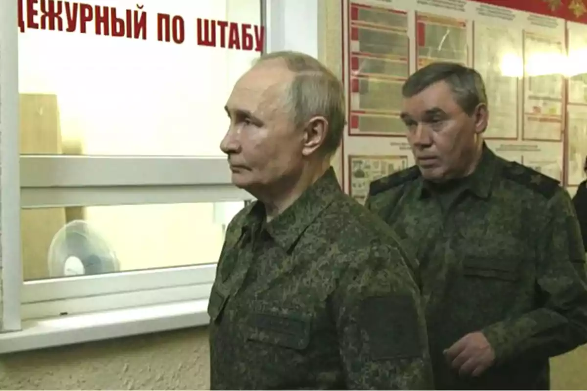 Two men in camouflage military uniforms are standing indoors, one behind the other, facing a window with a sign in Russian.