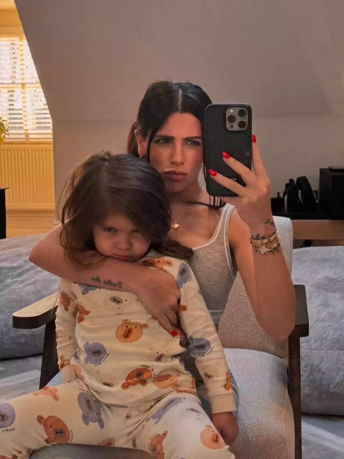 A person with dark hair takes a mirror selfie while holding a small child in bear pajamas, both with serious expressions. A person with dark hair takes a mirror selfie while holding a small child in bear pajamas, both with serious expressions.