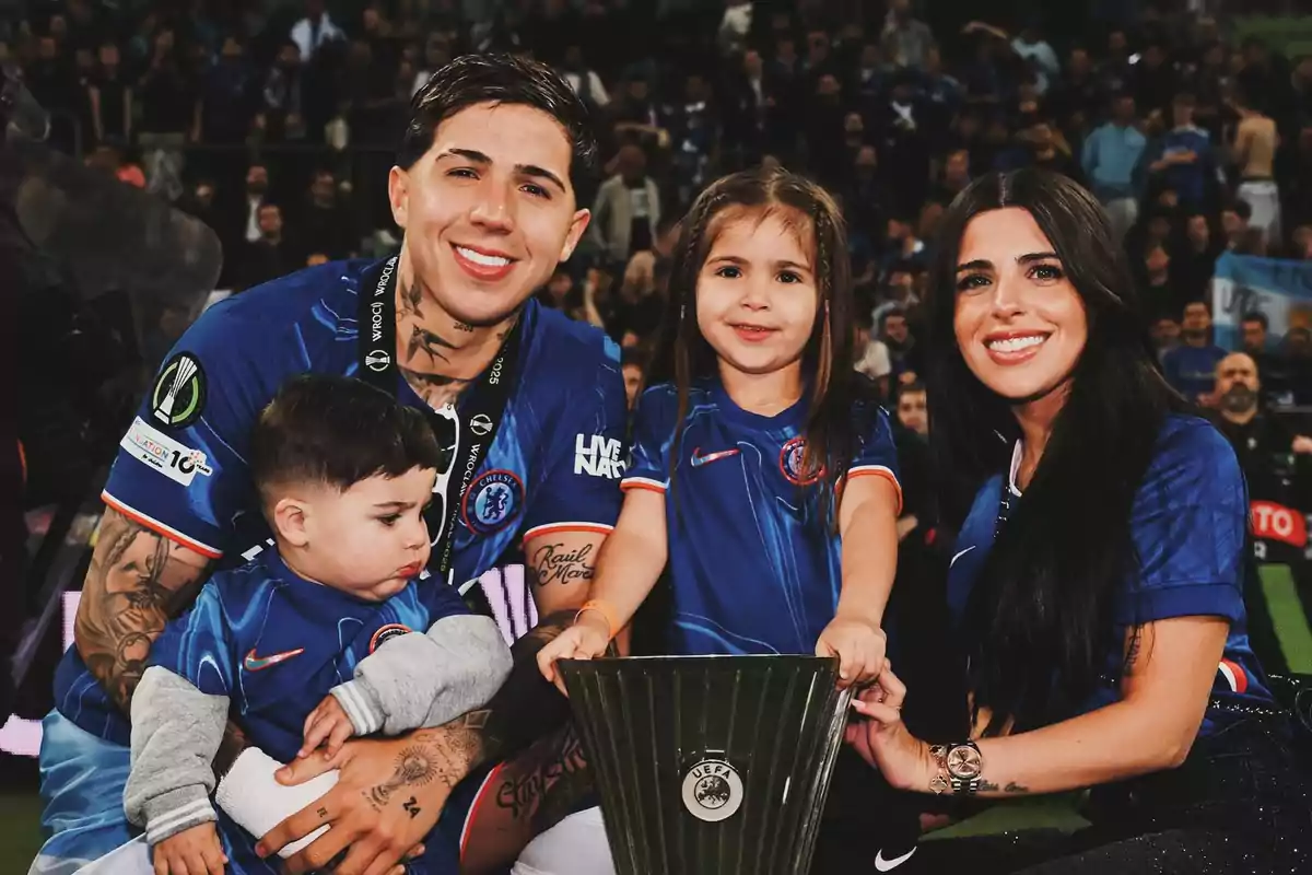 Una familia sonriente con camisetas de fútbol azul posa junto a un trofeo en un estadio lleno de gente Una familia sonriente con camisetas de fútbol azul posa junto a un trofeo en un estadio lleno de gente
