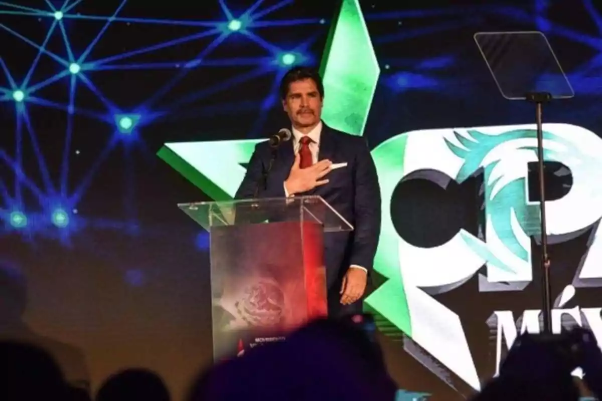 Hombre de traje azul y corbata roja hablando en un podio durante un evento con fondo de gráficos digitales y una estrella verde Hombre de traje azul y corbata roja hablando en un podio durante un evento con fondo de gráficos digitales y una estrella verde