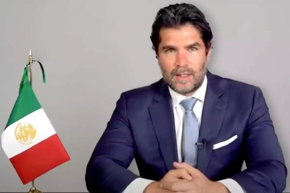 A man in a blue suit and gray tie is sitting in front of a Mexican flag against a gray background.