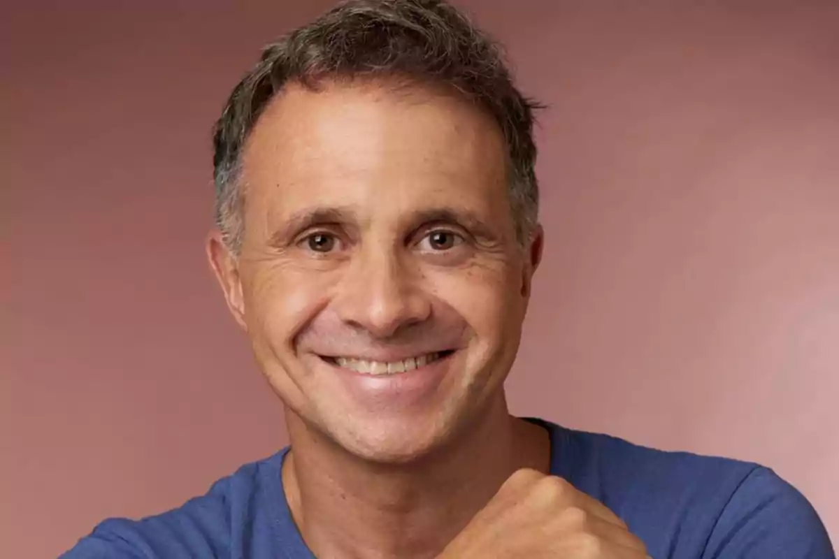 Un hombre sonriente con camiseta azul sobre un fondo marrón.