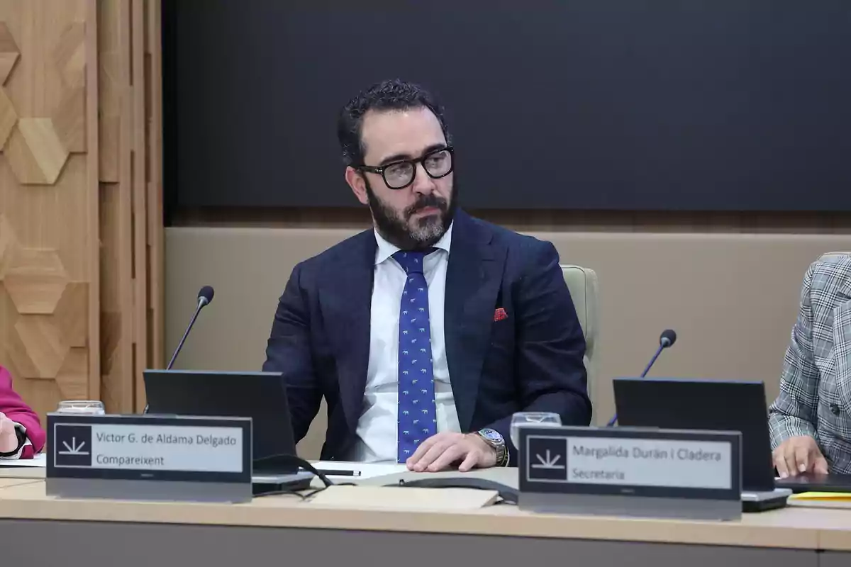 Un hombre con barba y gafas está sentado en una mesa de conferencias junto a otras personas, con un letrero frente a él.