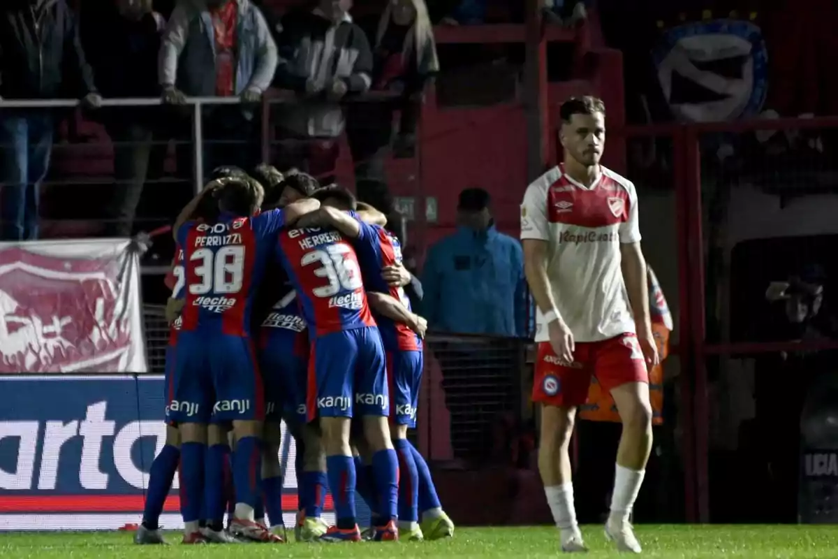 Players from a soccer team in blue and red uniforms celebrate in a group while a player from the opposing team in a white and red uniform walks near them on the field. Players from a soccer team in blue and red uniforms celebrate in a group while a player from the opposing team in a white and red uniform walks near them on the field.