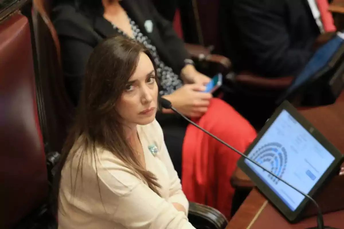 Una mujer sentada en un escritorio con un micrófono frente a ella y una tableta mostrando un gráfico, rodeada de otras personas en un entorno formal.