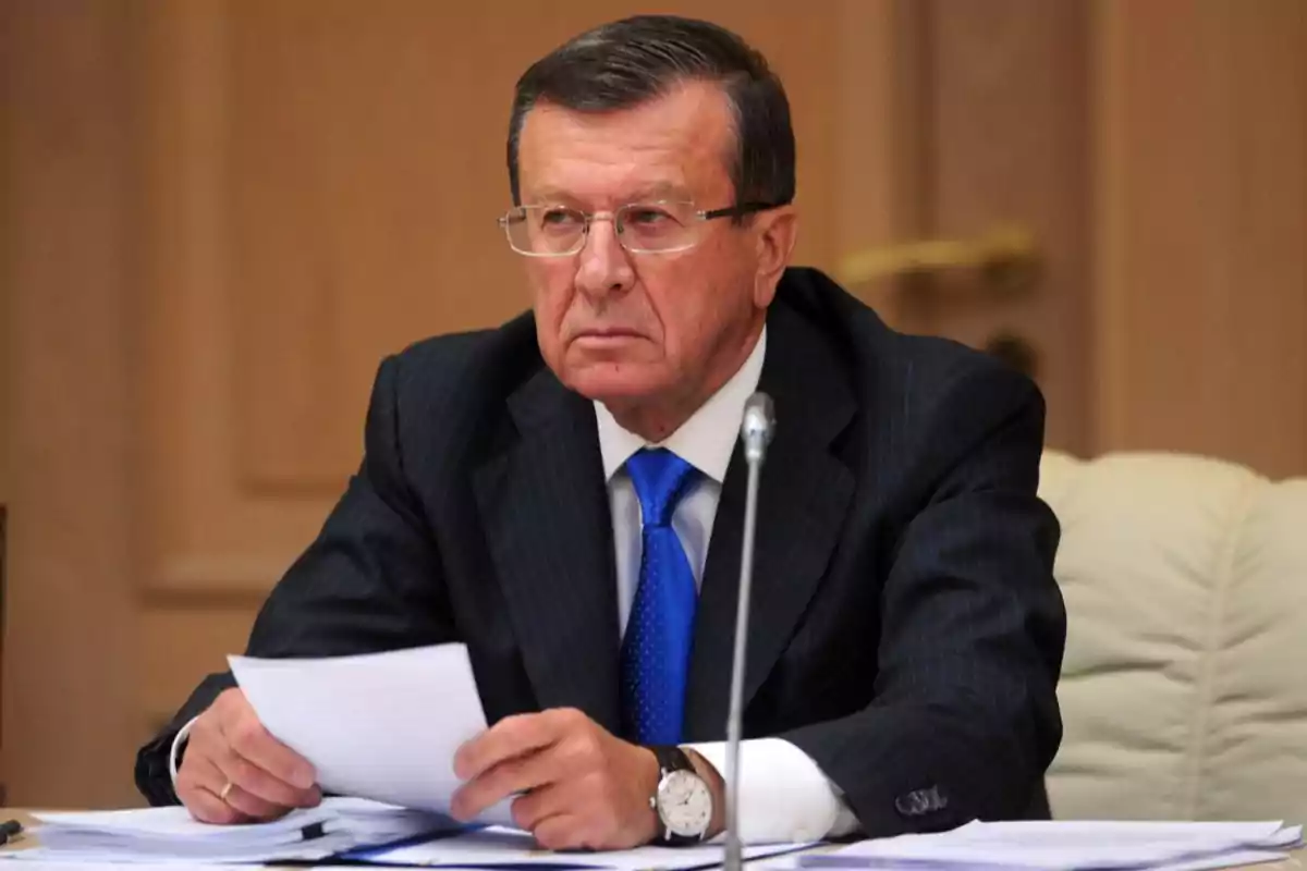 Un hombre de mediana edad con gafas y traje oscuro sostiene papeles mientras está sentado en una mesa, con una expresión seria y concentrada. Un hombre de mediana edad con gafas y traje oscuro sostiene papeles mientras está sentado en una mesa, con una expresión seria y concentrada.