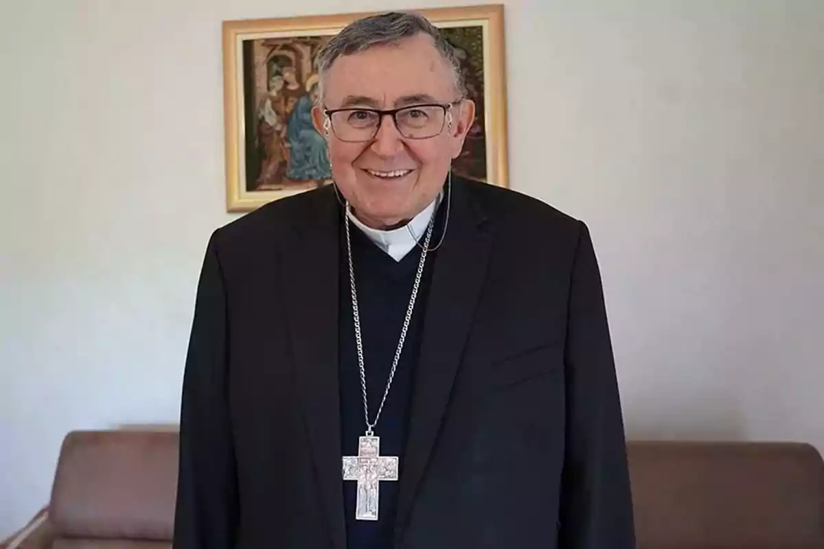 An elderly man with glasses and religious attire smiles in front of a painting on the wall. An elderly man with glasses and religious attire smiles in front of a painting on the wall.