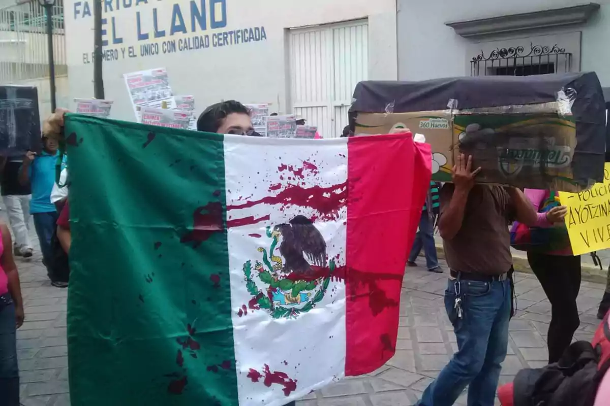 Una persona sostiene una bandera de México con manchas rojas en una manifestación en la calle, rodeada de otras personas que llevan carteles y cajas.