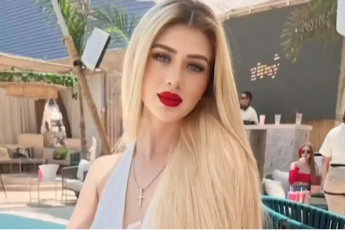 A person with blonde hair and red lips is in an outdoor setting with tropical decor and people in the background.