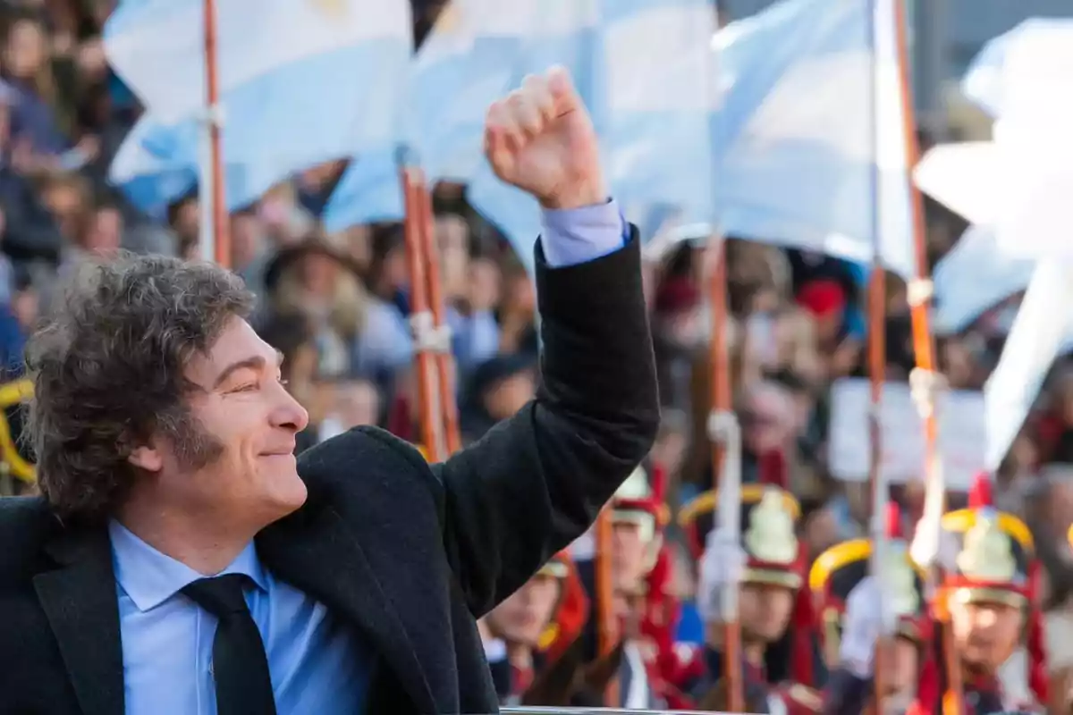 Un hombre sonriente con el puño levantado en un evento público con banderas argentinas de fondo.