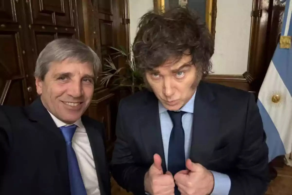 Dos hombres en traje posan para una foto en un ambiente formal con una bandera argentina al fondo.