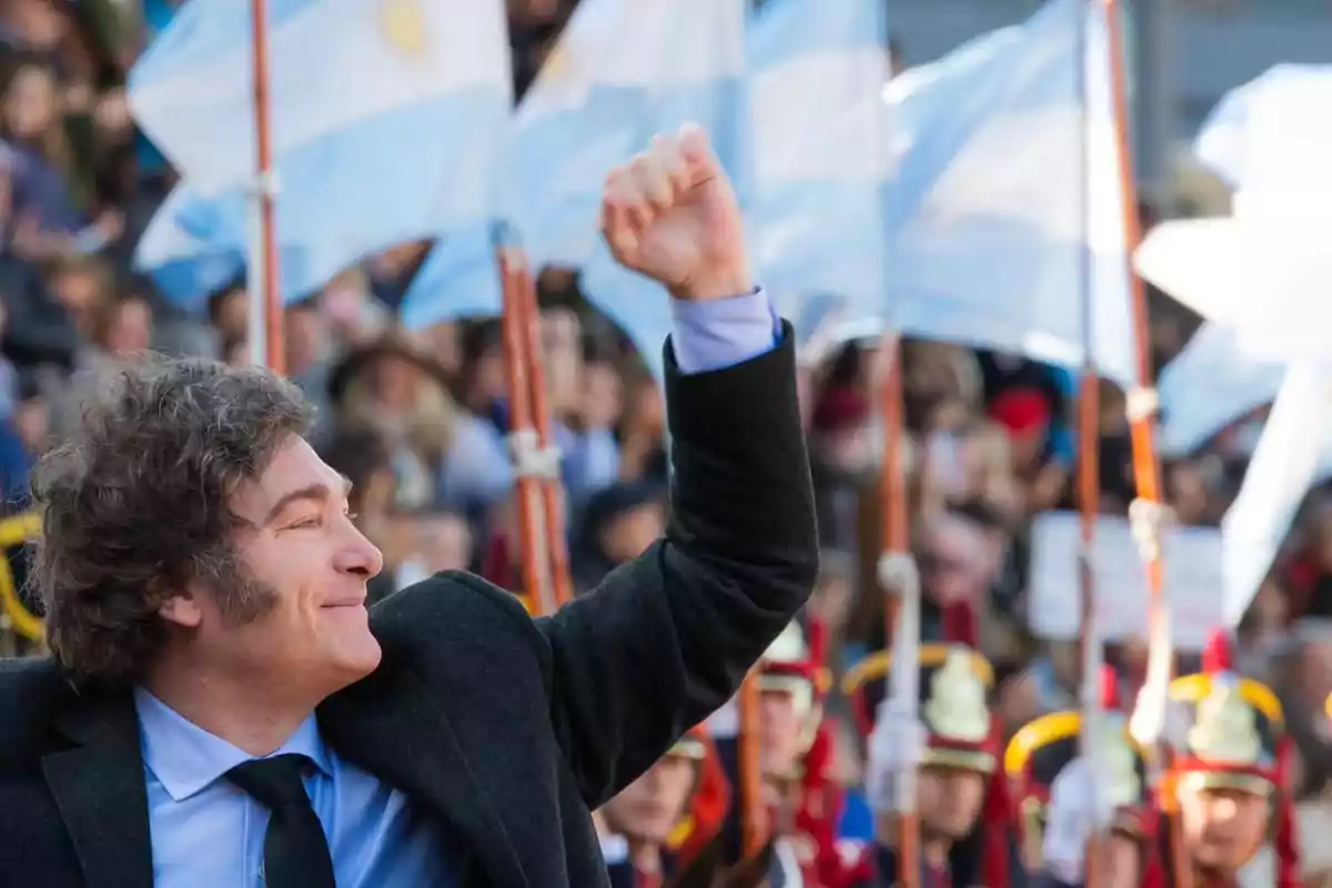 Un hombre sonriente levanta el puño en un evento al aire libre con banderas argentinas ondeando de fondo. Un hombre sonriente levanta el puño en un evento al aire libre con banderas argentinas ondeando de fondo.