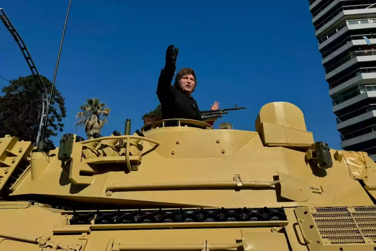 Persona saludando desde la escotilla de un tanque militar amarillo en una ciudad con cielo despejado y edificios modernos al fondo