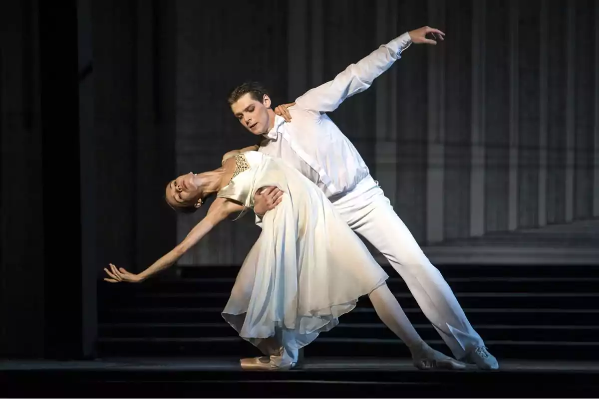 Una pareja de bailarines de ballet realiza una elegante pose en el escenario con el fondo oscuro.