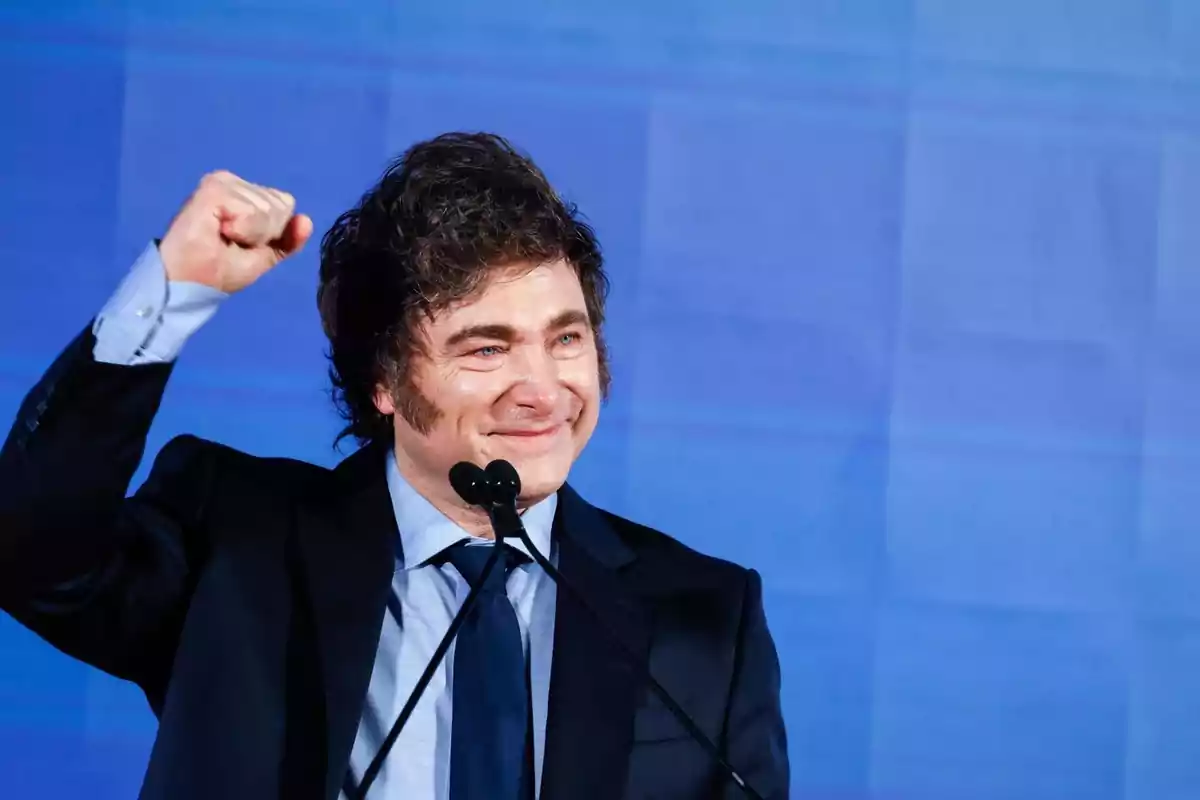 Un hombre sonriente con traje y corbata levanta el puño en señal de celebración frente a un fondo azul.