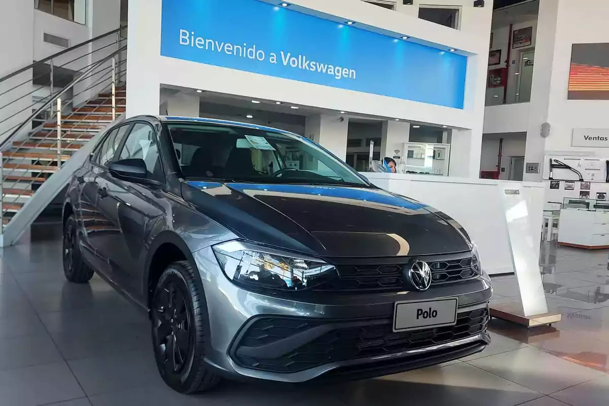 A dark gray Volkswagen Polo car is displayed inside a dealership with a sign that says "Welcome to Volkswagen" in the background.