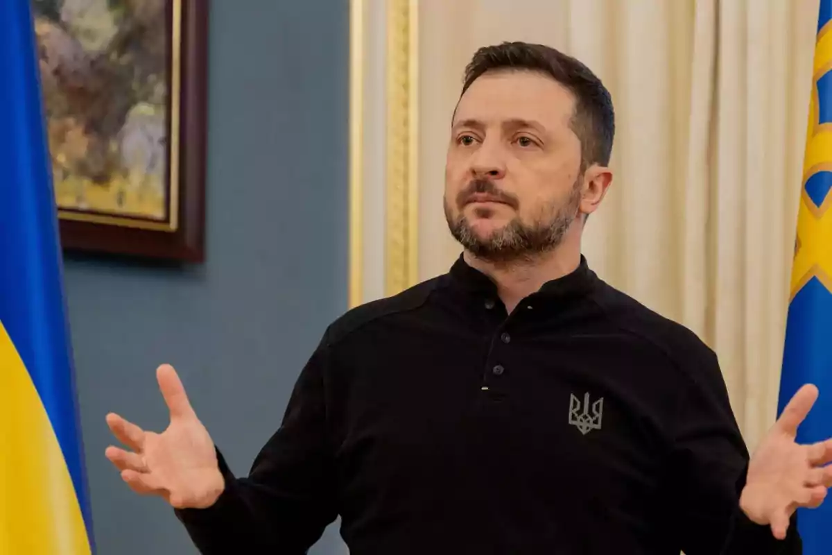 A man with a beard and short hair, wearing a black jersey, gestures with open hands in an indoor setting with a flag and a painting on the wall. A man with a beard and short hair, wearing a black jersey, gestures with open hands in an indoor setting with a flag and a painting on the wall.
