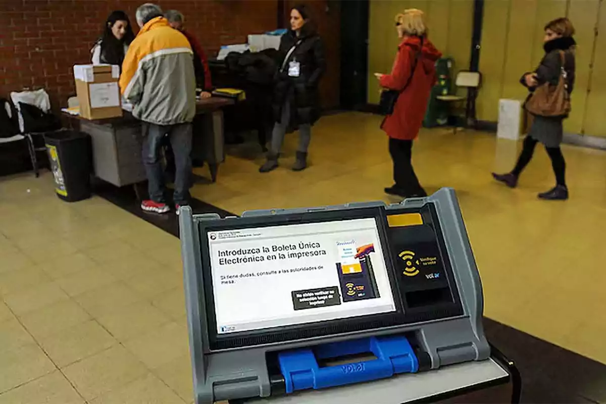 Personas en un centro de votación con una máquina de votación electrónica en primer plano. Personas en un centro de votación con una máquina de votación electrónica en primer plano.