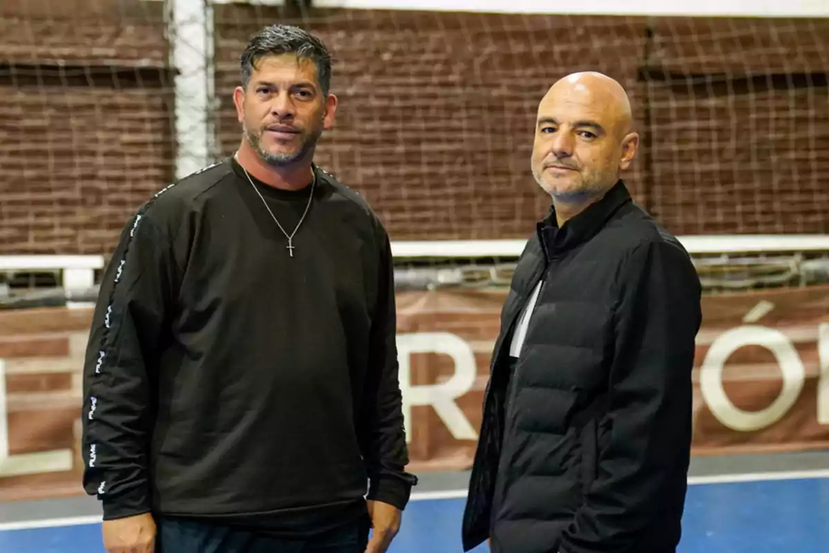 Dos hombres posan en una cancha deportiva cubierta, uno lleva un suéter negro y collar con cruz, el otro viste una chaqueta negra y camiseta clara. Dos hombres posan en una cancha deportiva cubierta, uno lleva un suéter negro y collar con cruz, el otro viste una chaqueta negra y camiseta clara.