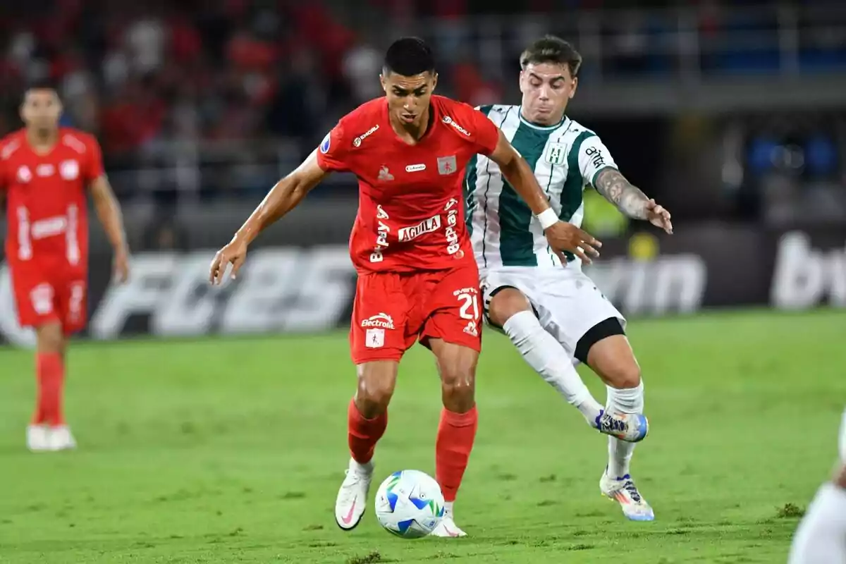 Two players compete for the ball during a match, one wearing a red uniform and the other wearing a white and green uniform, in a soccer stadium. Two players compete for the ball during a match, one wearing a red uniform and the other wearing a white and green uniform, in a soccer stadium.