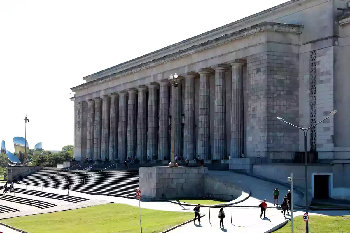 Fachada de un edificio con columnas grandes y personas caminando por la explanada en un día soleado