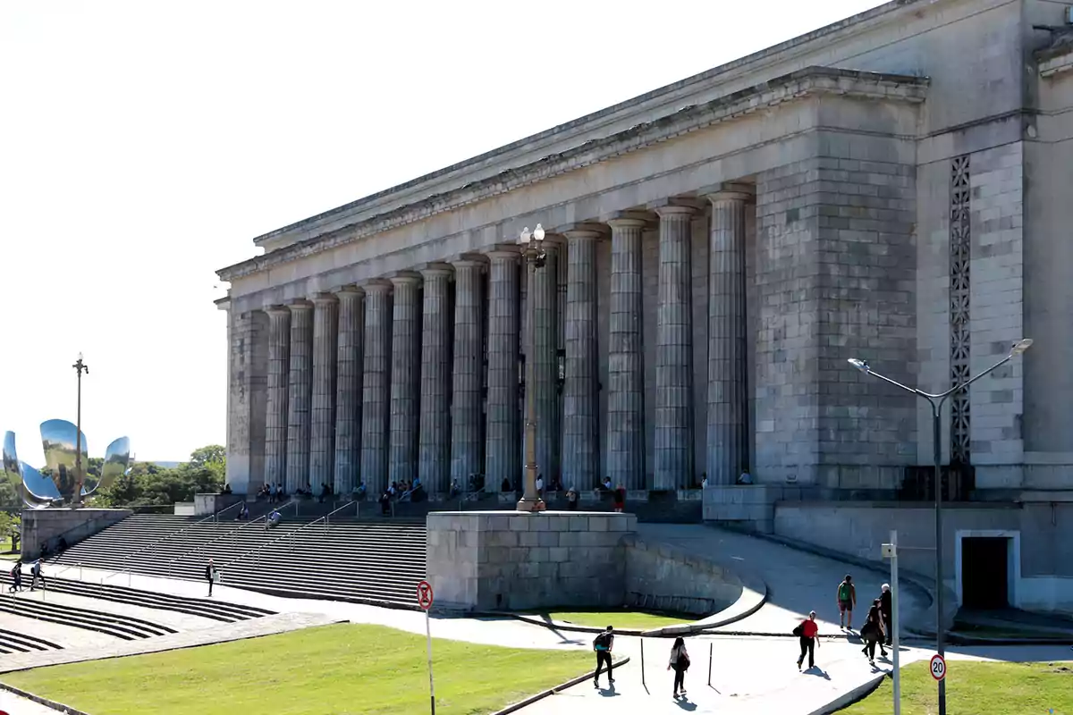 Facade of a neoclassical-style building with large columns and staircases, people walking, and a metal sculpture in the background surrounded by green areas