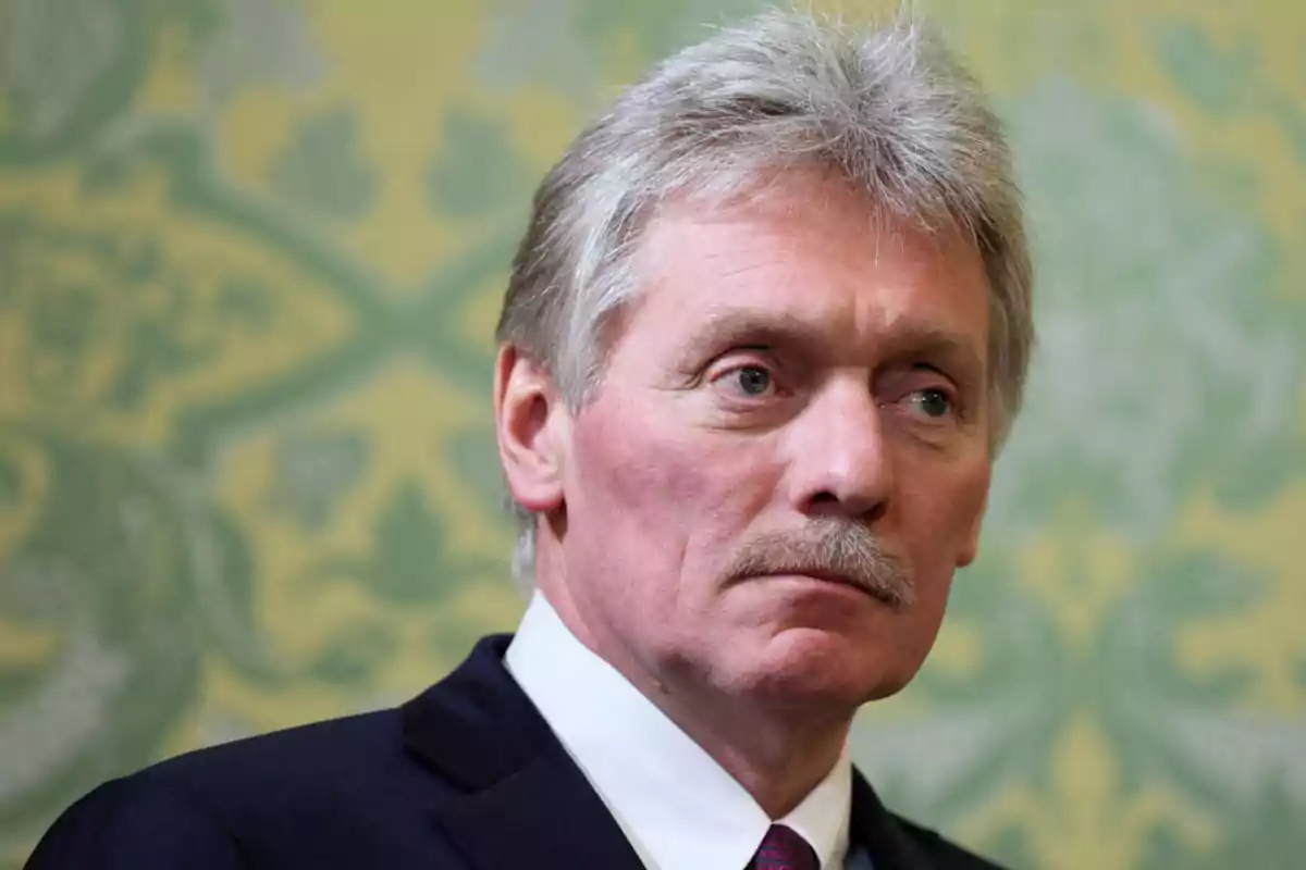Gray-haired man with a mustache and a serious expression, wearing a dark suit and tie, in front of a green and yellow decorative background Gray-haired man with a mustache and a serious expression, wearing a dark suit and tie, in front of a green and yellow decorative background