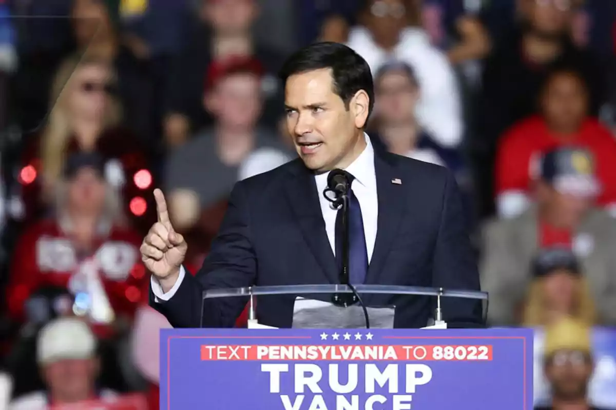 Hombre de traje oscuro hablando en un mitin político con un letrero que dice Trump Vance frente a una multitud desenfocada Hombre de traje oscuro hablando en un mitin político con un letrero que dice Trump Vance frente a una multitud desenfocada