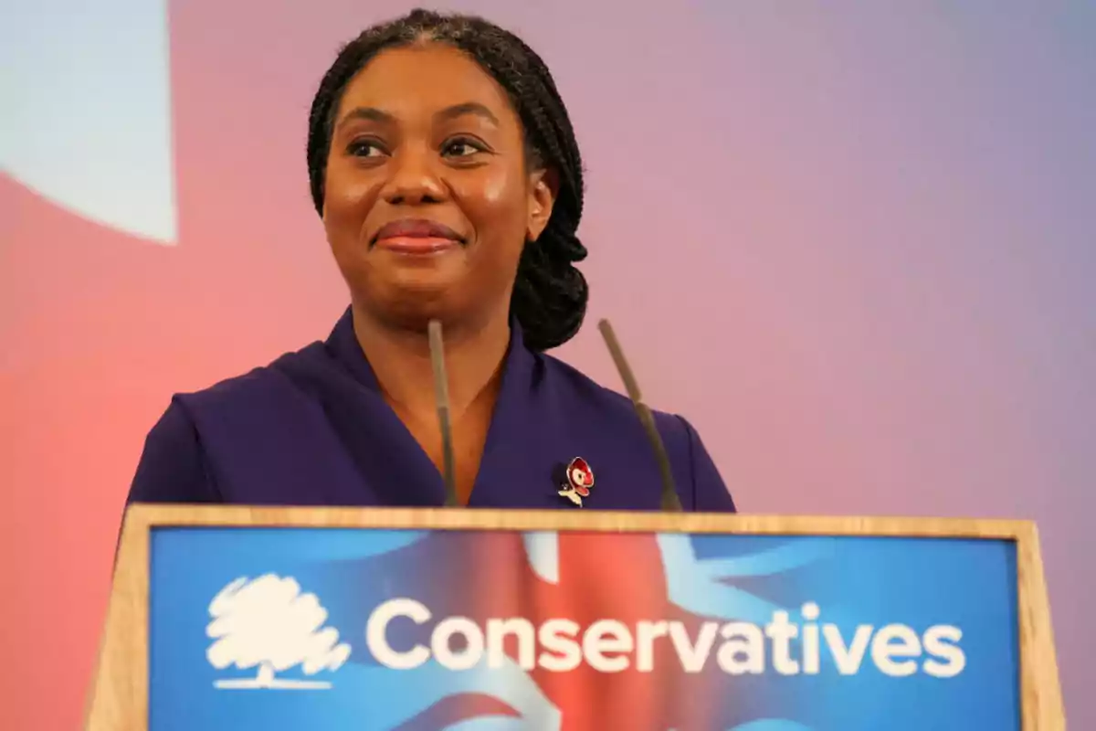 A person standing behind a podium with the conservative party logo, smiling and looking to the side. A person standing behind a podium with the conservative party logo, smiling and looking to the side.