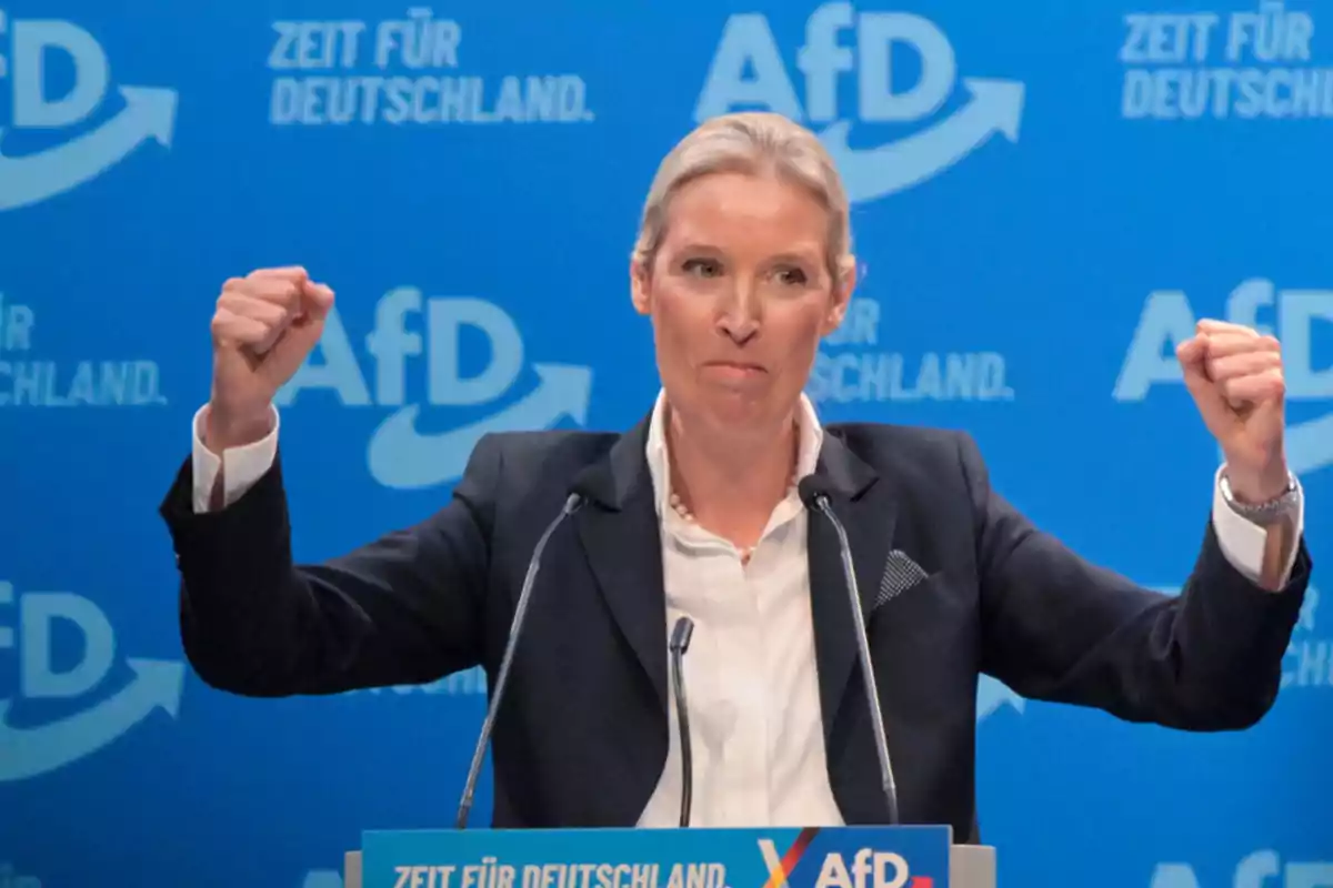 Una persona en un podio con fondo azul y logotipos de AfD levantando los puños. Una persona en un podio con fondo azul y logotipos de AfD levantando los puños.