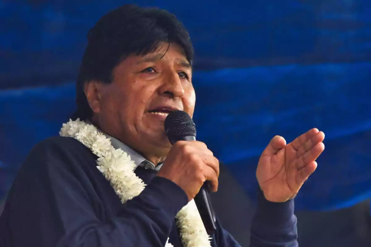 Hombre de cabello oscuro hablando por micrófono y usando un collar de flores blancas frente a un fondo azul Hombre de cabello oscuro hablando por micrófono y usando un collar de flores blancas frente a un fondo azul