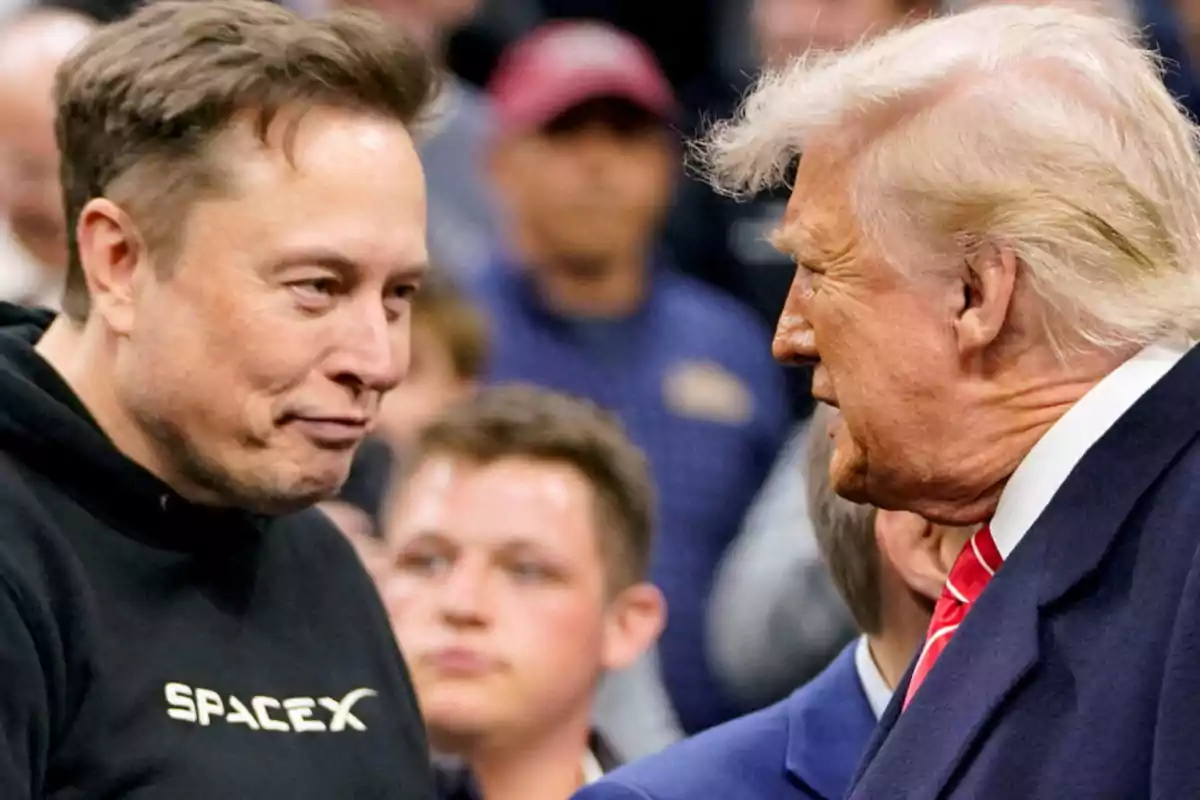 Two men are talking face to face at an event with several people in the background; one is wearing a black sweatjersey with the SpaceX logo and the other is dressed in a blue suit with a red tie.