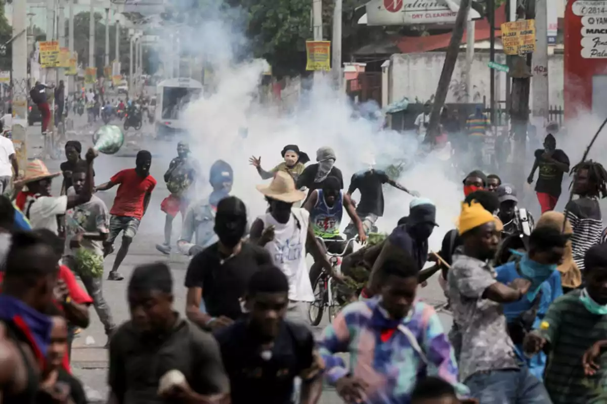 Personas corren y protestan en una calle llena de humo mientras algunas llevan el rostro cubierto y otras lanzan objetos