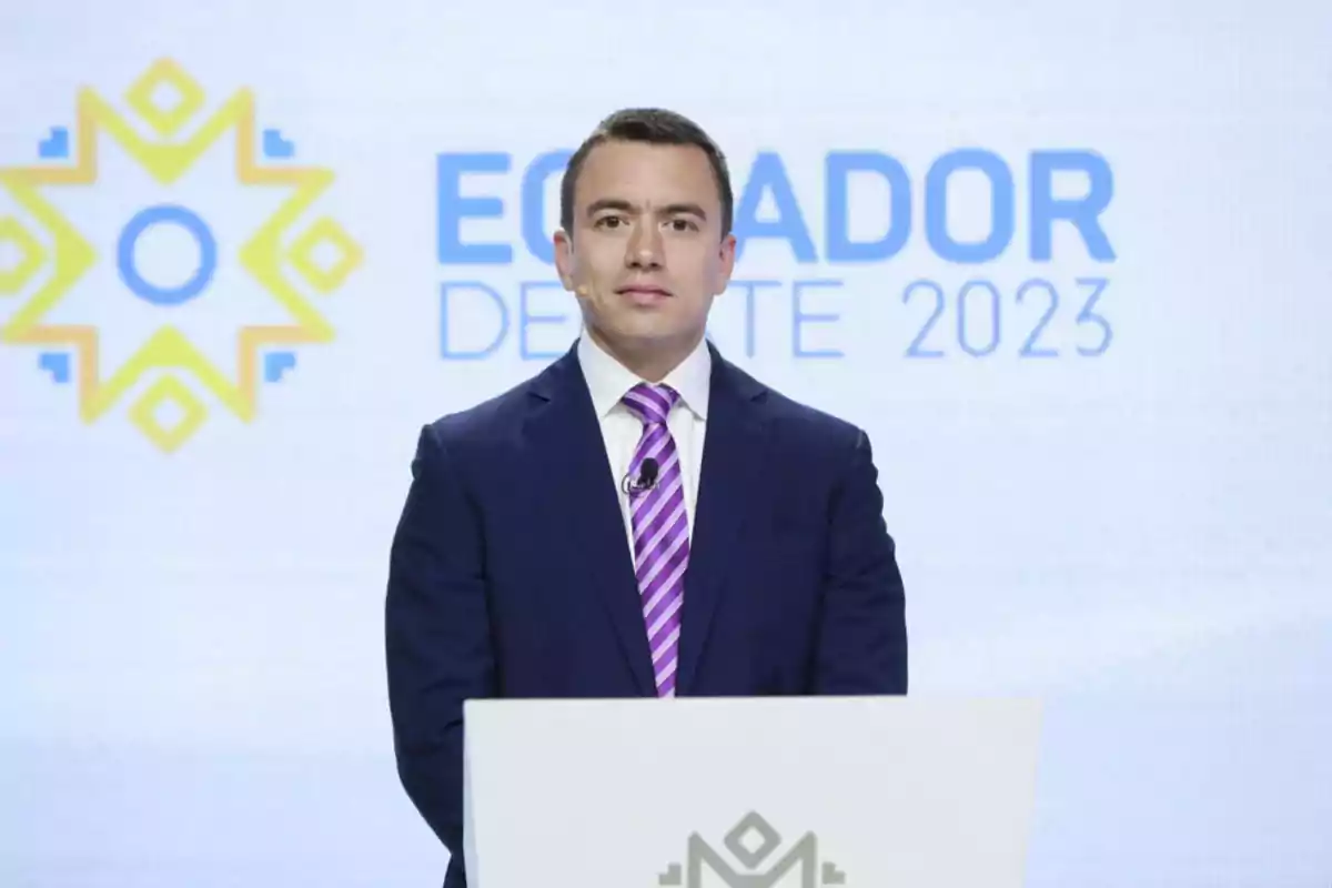 Un hombre de traje oscuro y corbata morada está de pie frente a un podio con un micrófono en un evento que tiene un fondo con el texto "Ecuador Debate 2023".